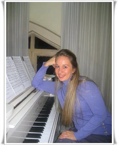 Ferzan in a blue top, smiling and leaning on a white upright piano
