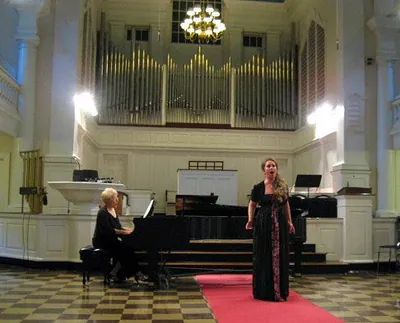 Ferzan singing on a red carpet in a white hall, with a pianist beside her and a pipe organ behind