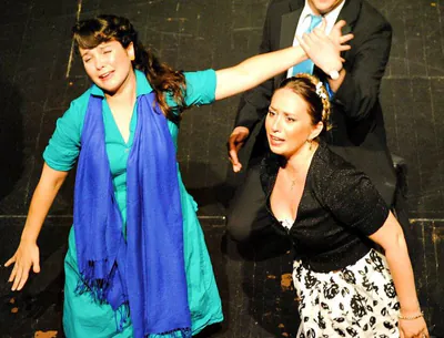 Two women in a dramatic stage scene with a man in a suit behind them